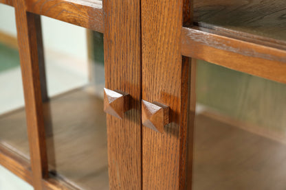 Close-up of a wooden cabinet or bookcase with two glass-front doors and matching geometric wooden knobs, showing warm medium-brown wood grain.