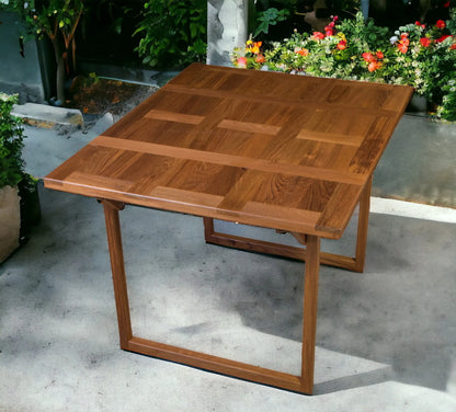 Wooden outdoor table with a checkered pattern on a concrete surface with plants in the background.