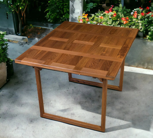 Wooden outdoor table with a checkered pattern on a concrete surface with plants in the background.