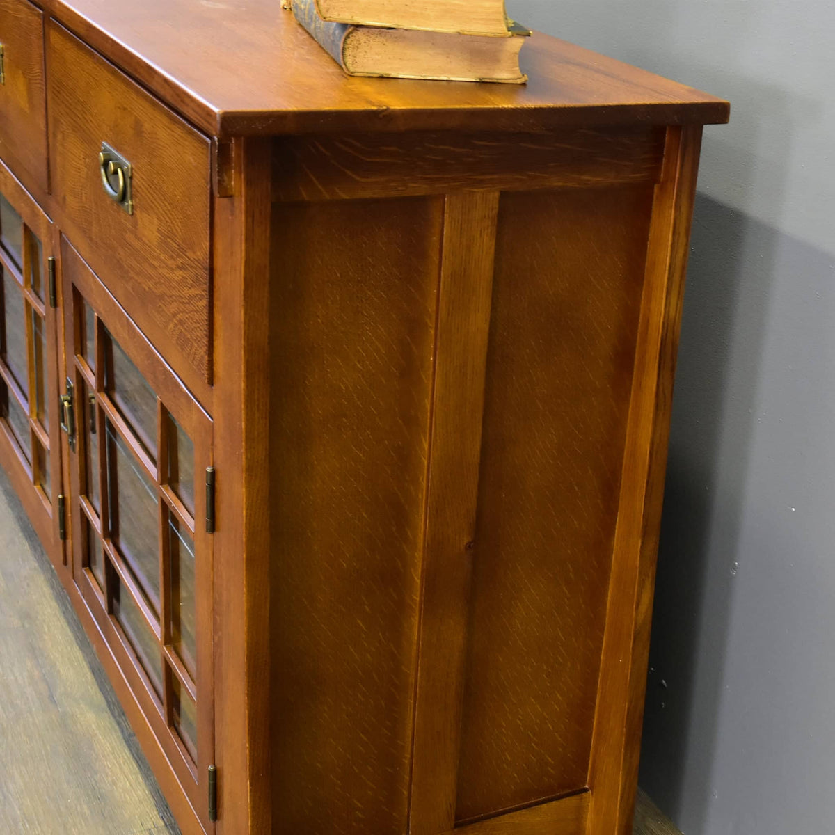 A warm brown wooden sideboard cabinet with a top drawer, two glass-paneled doors, brass hardware, and a stack of vintage books on top, photographed indoors against a gray wall.