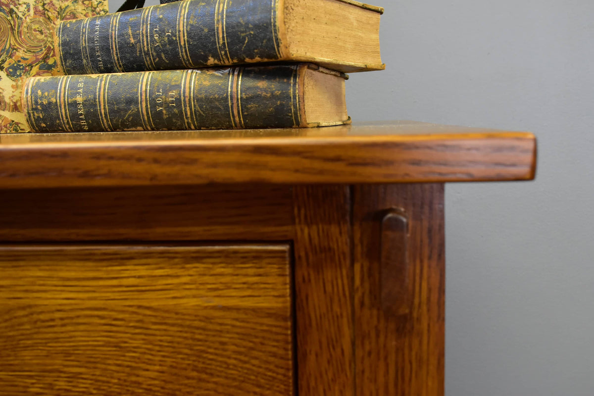 Close-up of a warm medium-brown wooden cabinet corner with a brass pull handle, holding a stack of antique dark blue books with gold lettering against a plain gray wall.