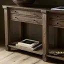 A rustic dark brown wooden sideboard or console table with three drawers, a lower open shelf holding a stack of papers, and a bowl on top, set against a light wood floor.