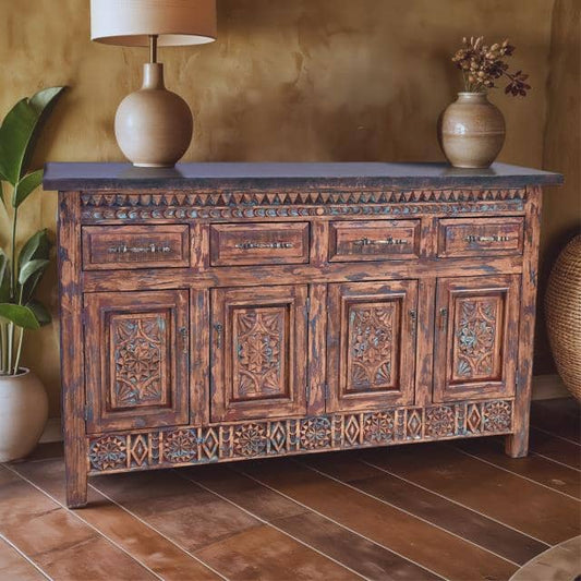 An ornate carved wooden sideboard or credenza with distressed brown and blue tones, topped with a dark blue surface holding a lamp and vase, set in a rustic room with a textured wall and wood floor.