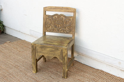 Wooden chair with intricate carvings on a textured floor against a white wall.