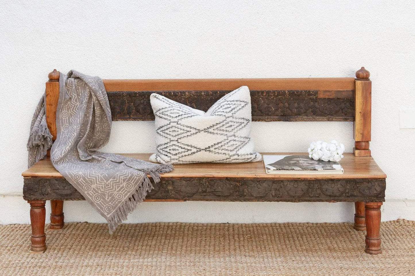 A rustic wooden bench with a carved dark brown backrest, styled with a gray patterned throw pillow, a draped gray throw blanket, a magazine, and a white decorative object, set against a white wall and jute rug.