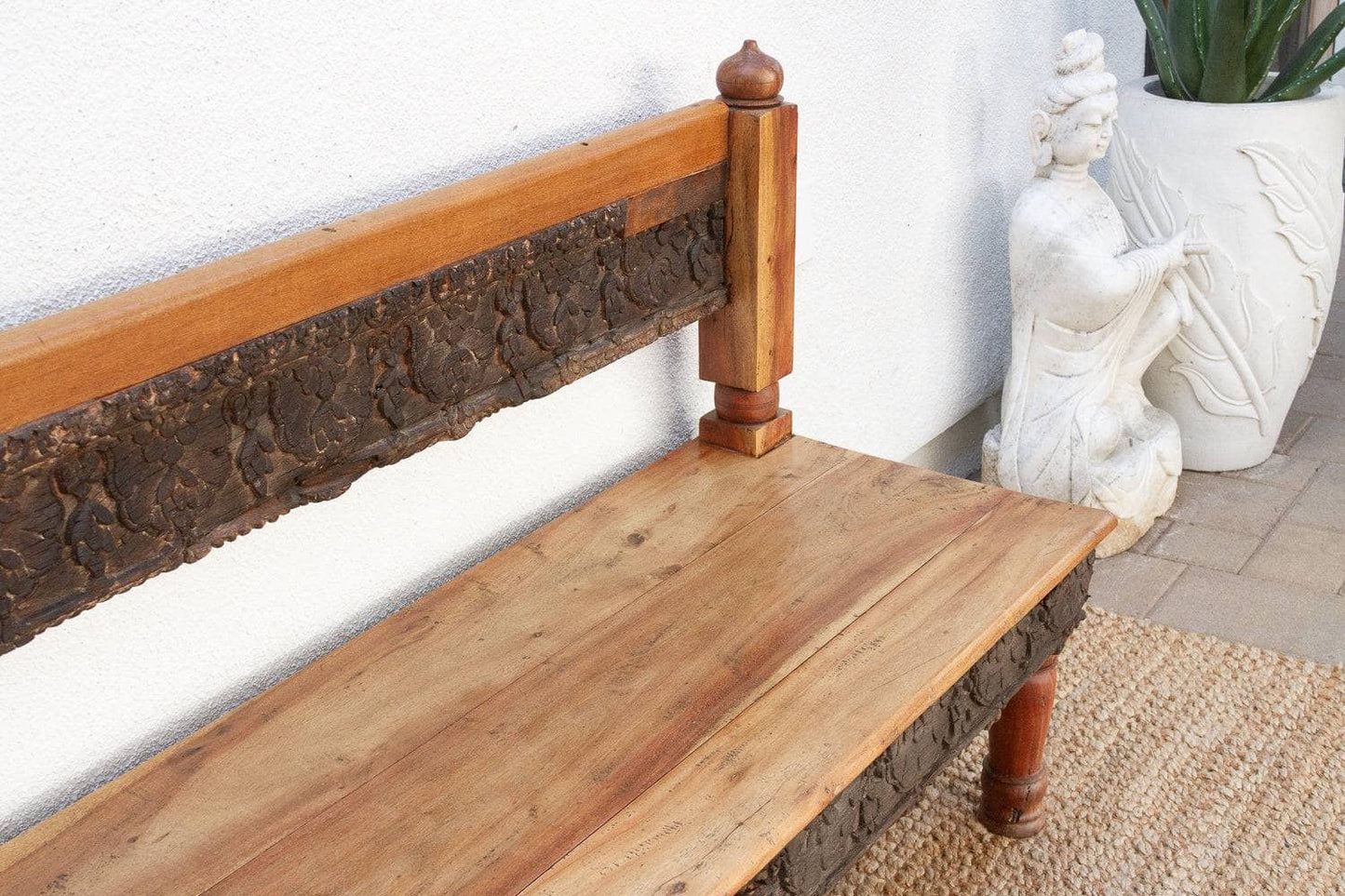 Wooden bench with rustic design against a white wall with a decorative statue and plant.