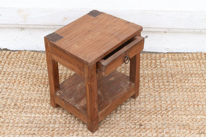 Wooden side table with a drawer on a textured floor.
